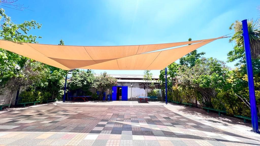 Vela de sombra color arena tensada sobre una plaza escolar de pastelones entre árboles