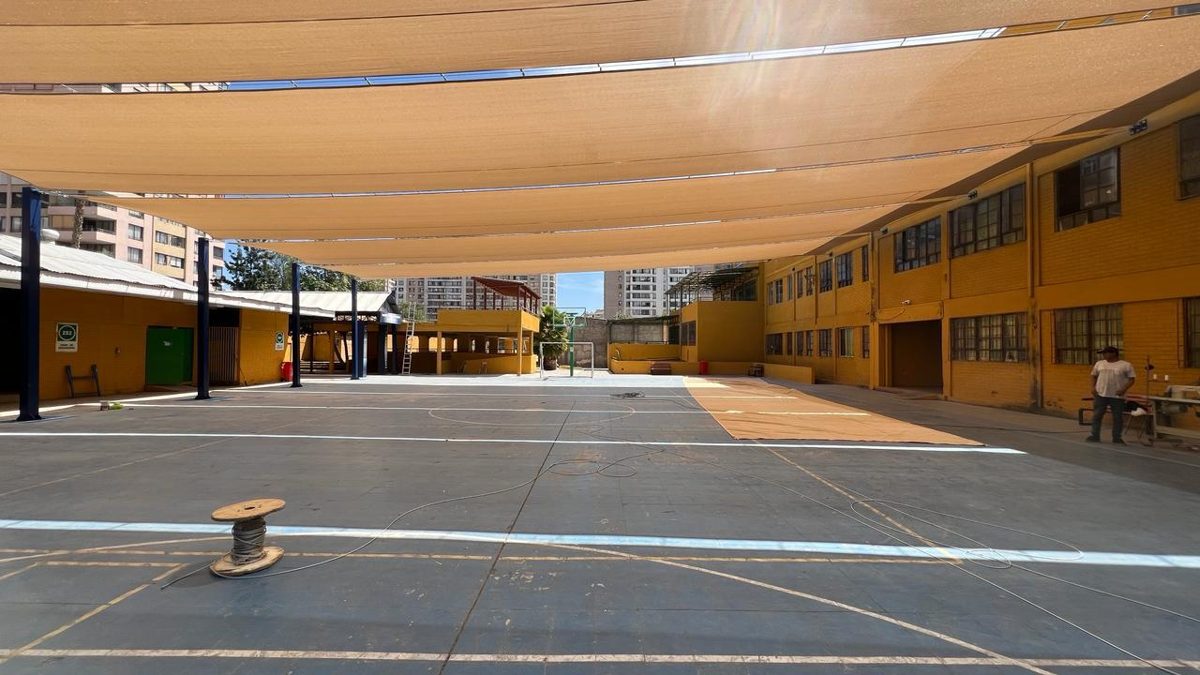 Velas de sombra color arena cubriendo el patio interior de un colegio de fachada amarilla
