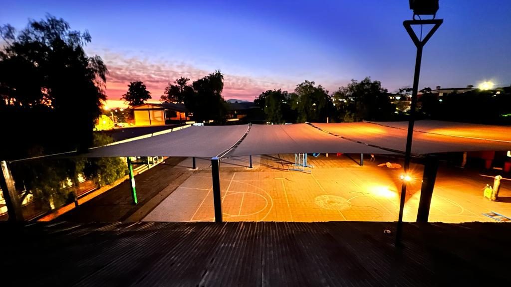 Velas de sombra sobre el patio de un colegio al atardecer con la cancha iluminada